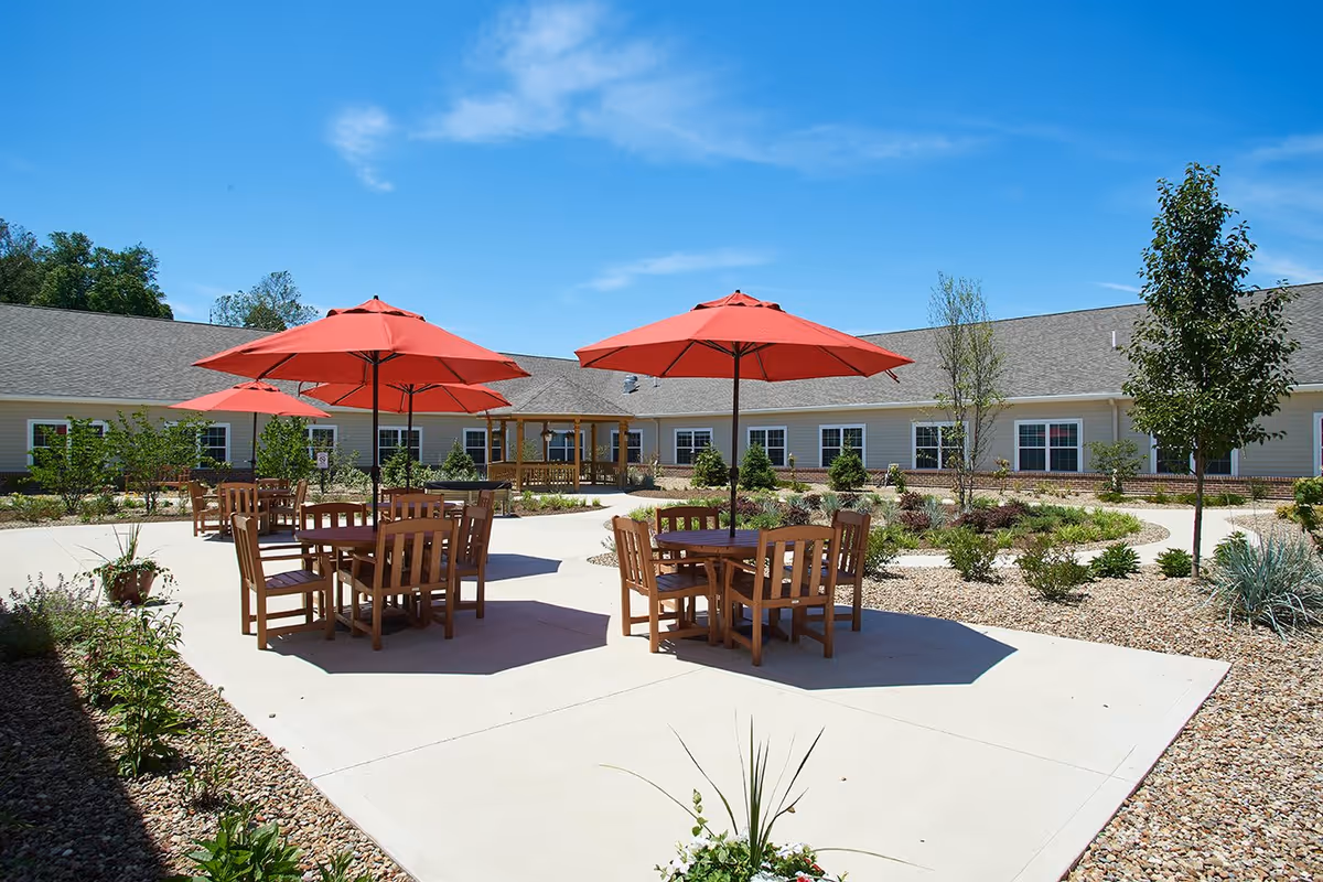 Outdoor courtyard area at Danbury Millersburg with wooden tables and chairs under red umbrellas, surrounded by landscaped plants and a building with multiple windows under a clear blue sky.