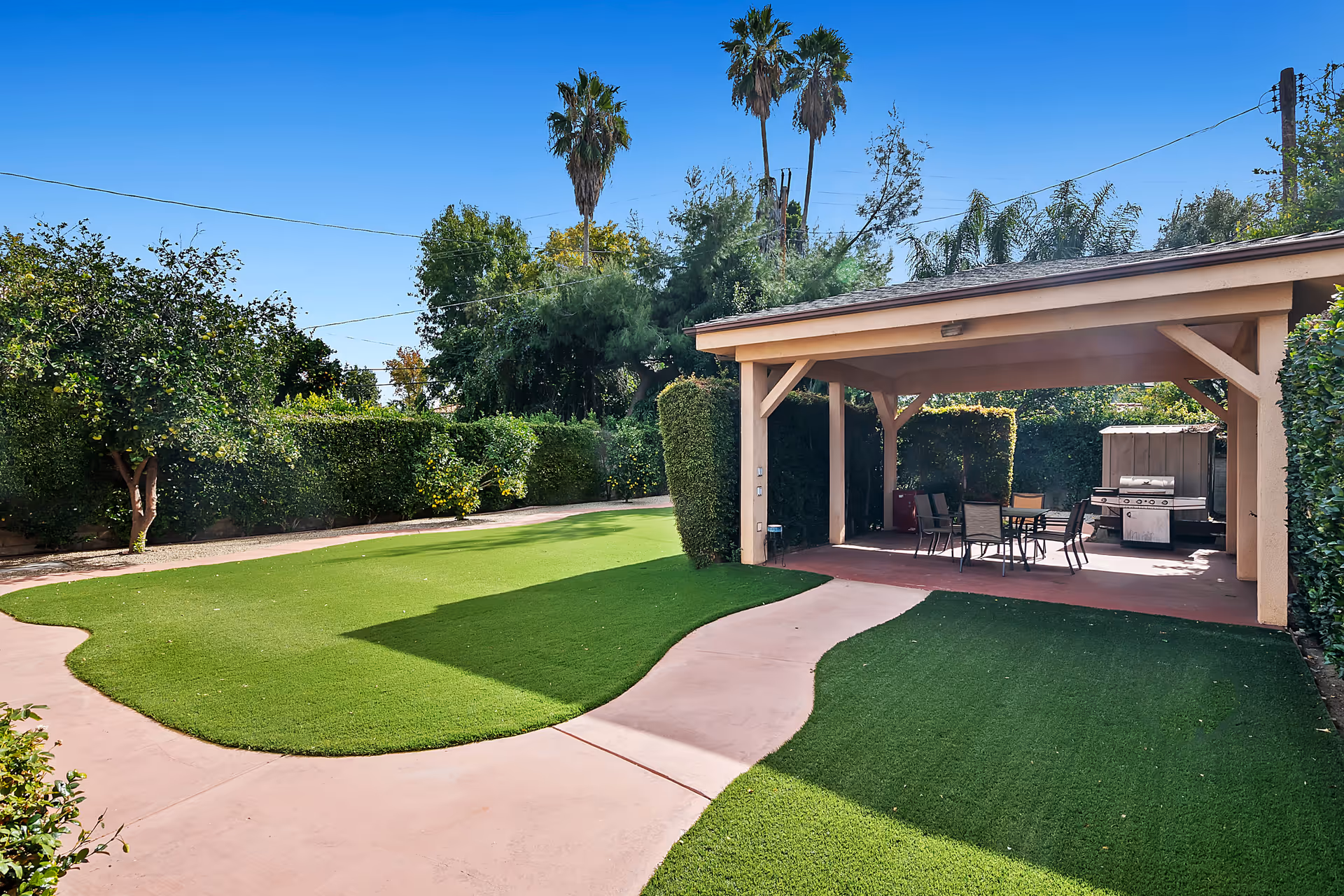 Outdoor patio area with a covered seating space featuring a table and chairs, a barbecue grill, and artificial grass lawn surrounded by trees and bushes under a clear blue sky.