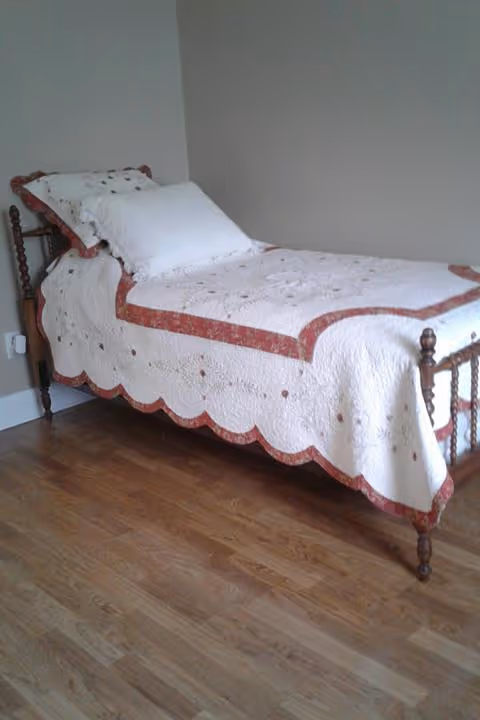 Single wooden twin bed with a white and red quilt against a beige wall on a hardwood floor.