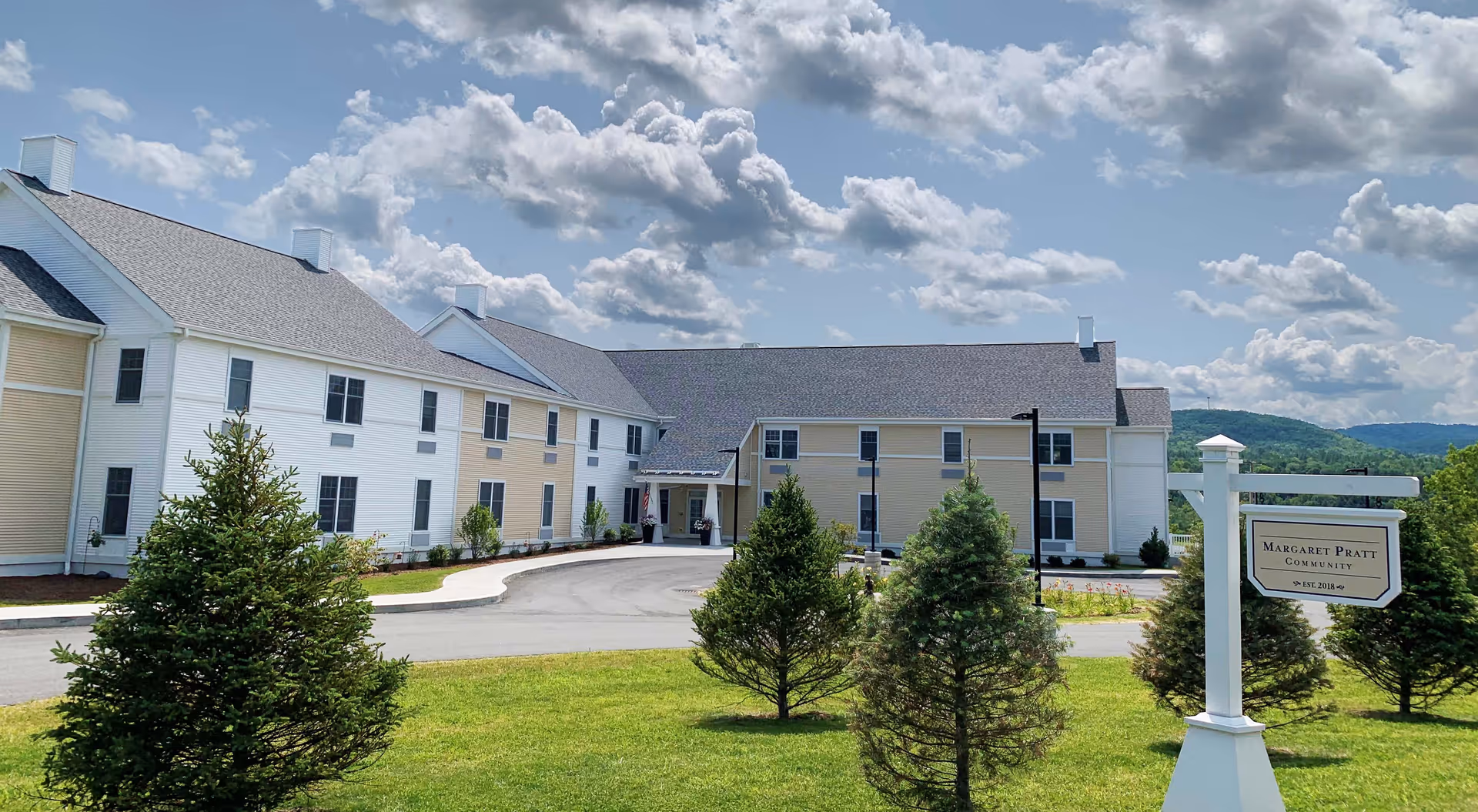 Exterior view of the Margaret Pratt Community building with a driveway, green lawn, several small pine trees, and a partly cloudy sky.