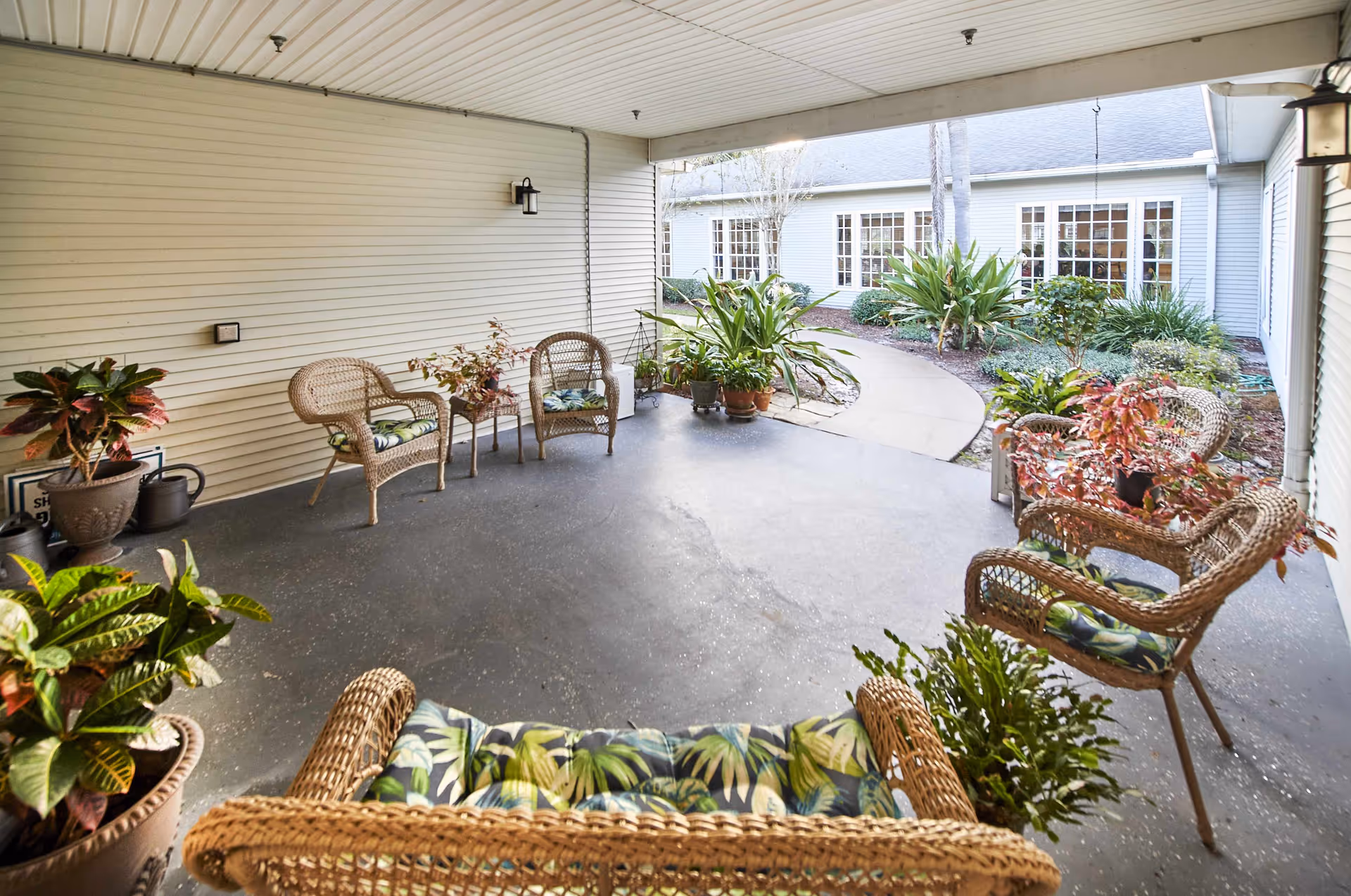 Covered patio area with wicker chairs and small tables, decorated with potted plants. The patio overlooks a garden path leading to a building with large windows.