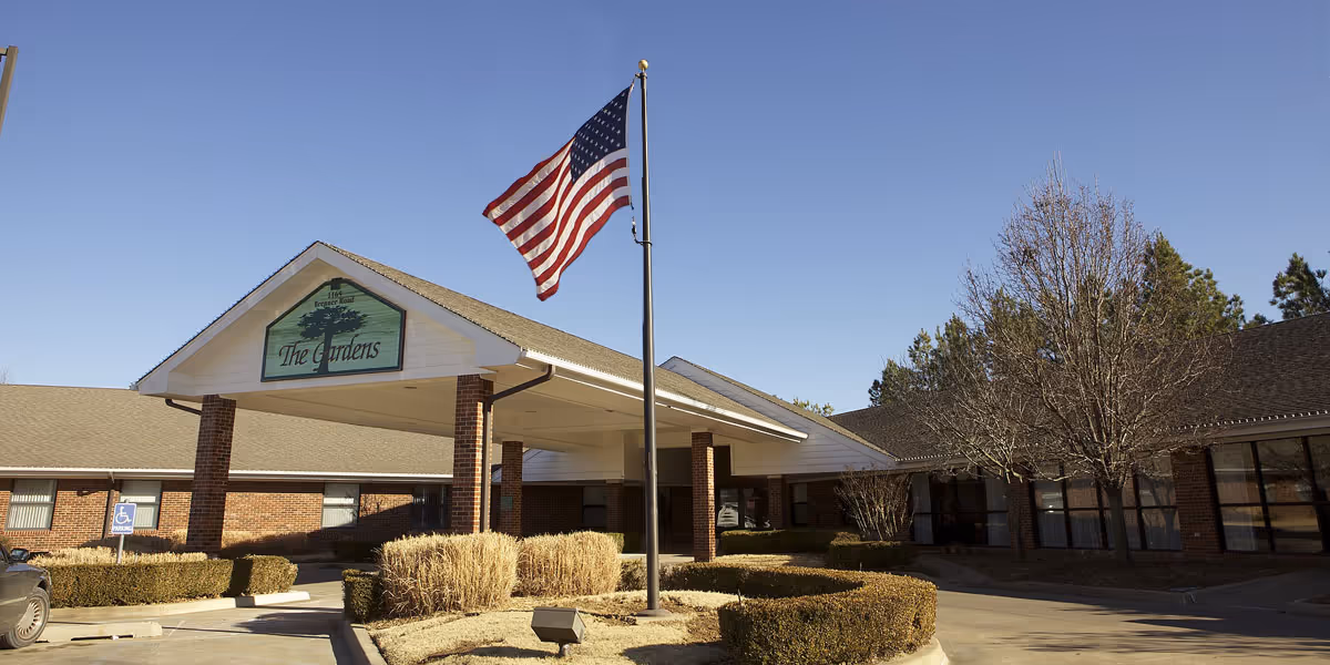 Exterior view of The Gardens senior living facility with a covered entrance supported by brick columns, an American flag on a flagpole in front, trimmed bushes, leafless trees, and a clear blue sky.