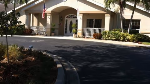 Front exterior view of a single-story building with a covered entrance, two American flags, and a driveway surrounded by landscaping with bushes and palm trees.