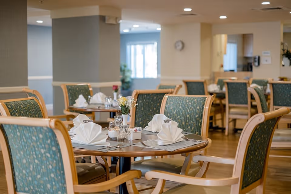 A dining room in a senior living facility with round wooden tables set with folded white napkins, silverware, salt and pepper shakers, and small flower vases. The chairs have wooden frames with green patterned upholstery. The room has soft lighting and a warm, inviting atmosphere.