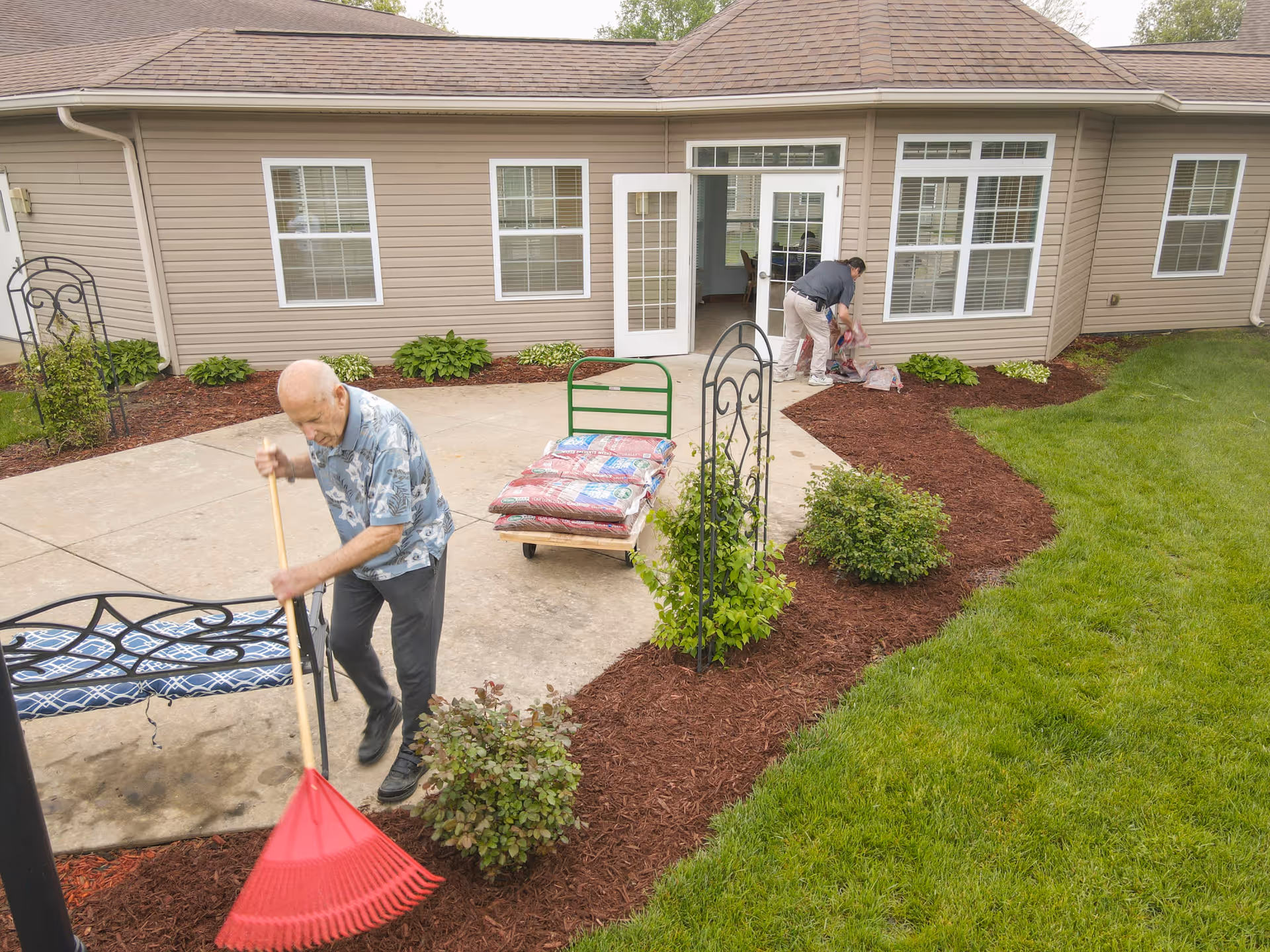 An elderly man raking mulch in a garden area outside a senior living facility building, while another person is arranging bags of mulch near the building entrance. The building has beige siding, multiple windows, and a glass door leading outside to a concrete patio with garden beds and green grass.