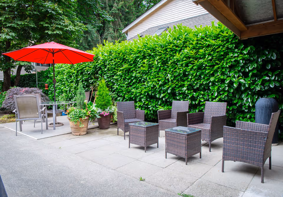 Outdoor patio area with several brown wicker chairs and matching small tables arranged on a paved surface. There is a red umbrella shading a small table with two chairs on the left side. Lush green bushes and trees provide a natural backdrop.