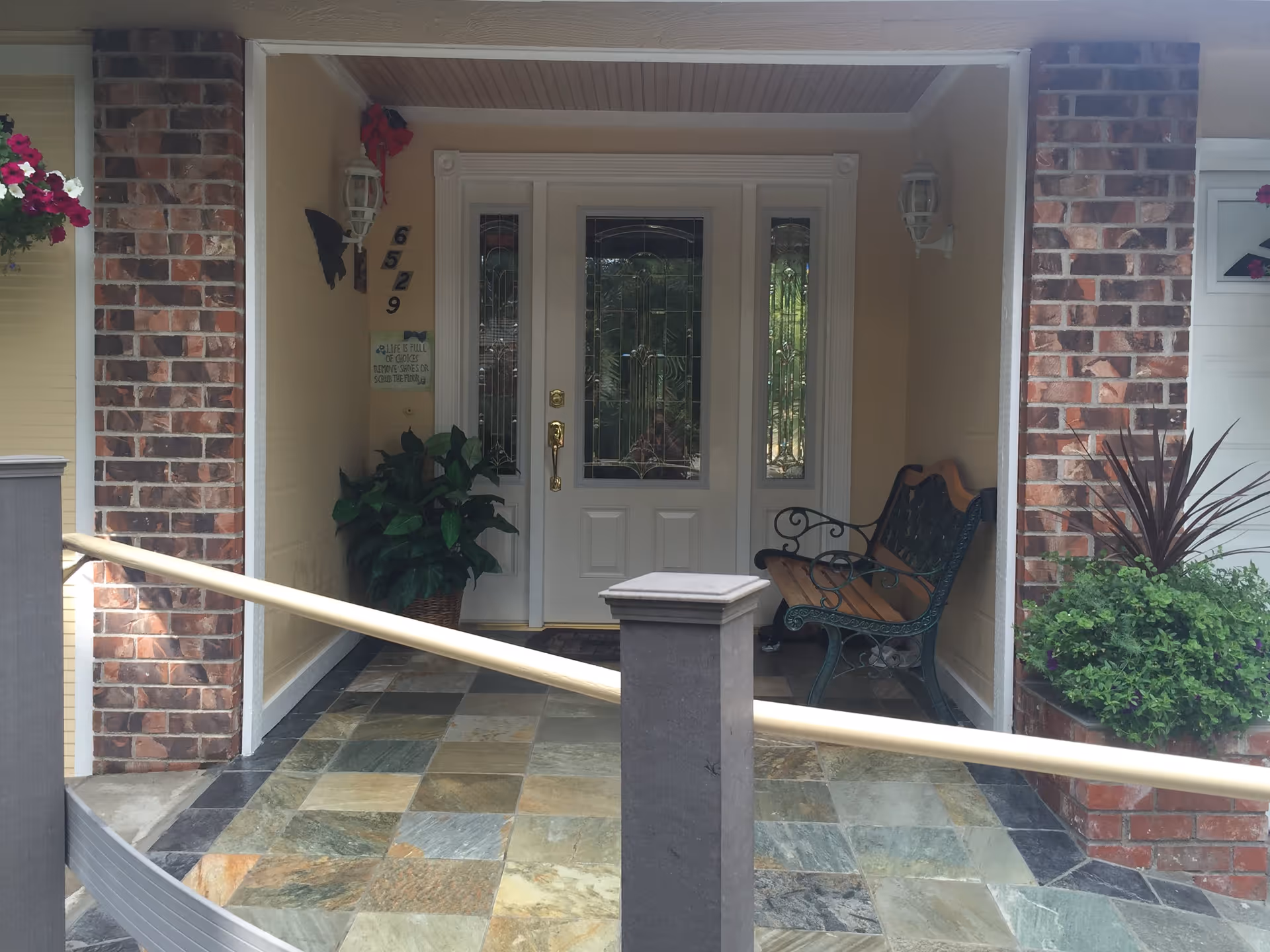 Front porch entrance of a house with a white door featuring decorative glass panels, a bench with wooden seat and ornate metal armrests, potted plants on either side, brick walls, and a tiled floor. There is a handrail leading up to the porch.