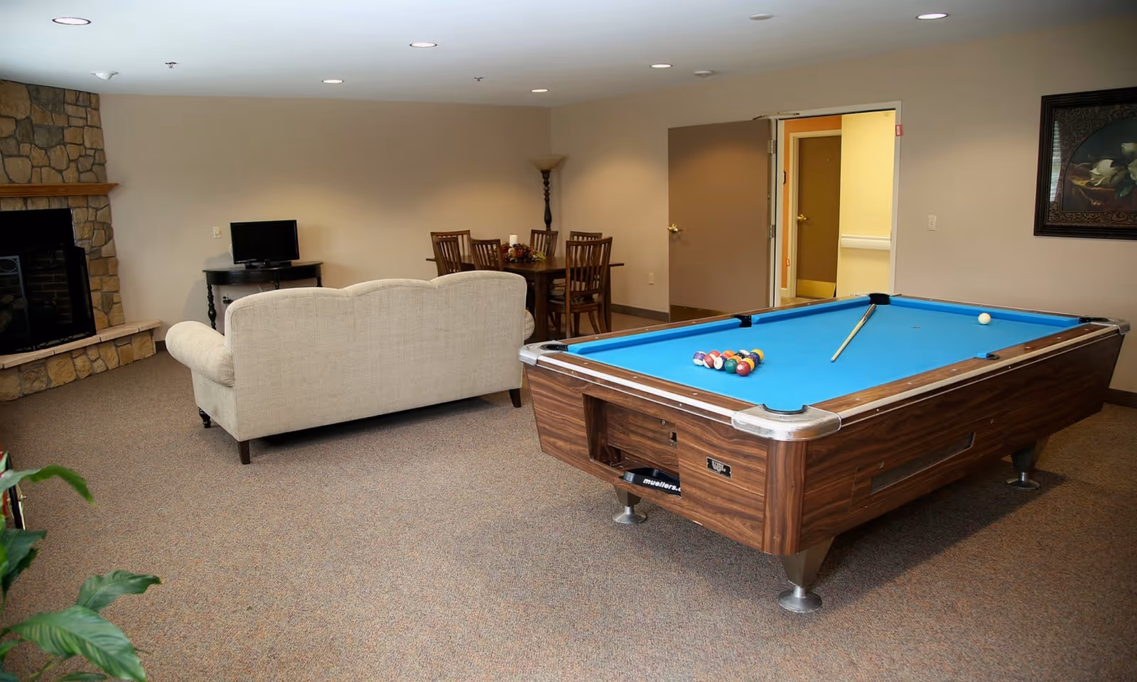 A recreational room with a pool table in the foreground, a beige couch facing a small TV on a black stand, a stone fireplace on the left wall, a wooden dining table with chairs in the background, and a framed floral painting on the right wall.