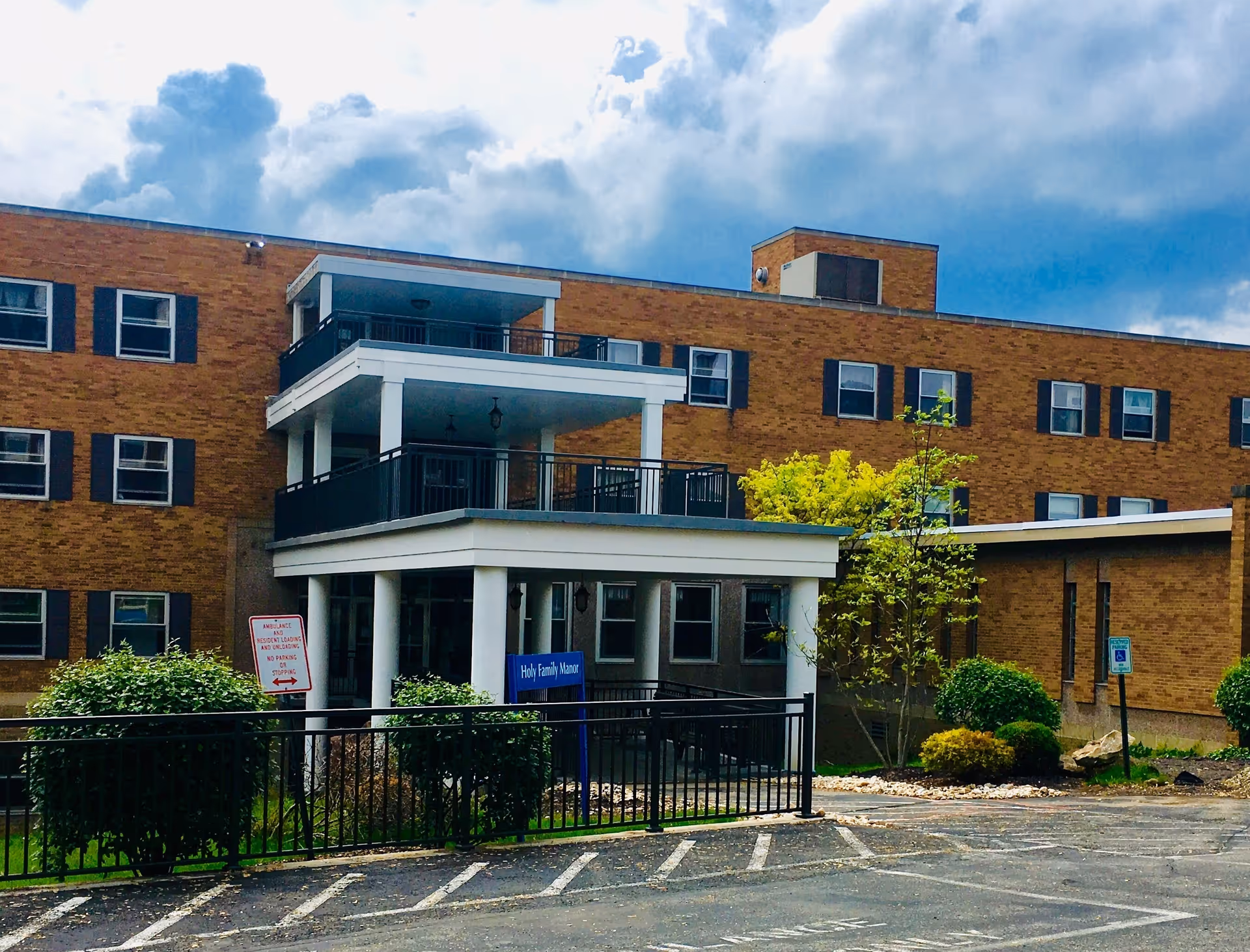Exterior view of a multi-story brick building with black window shutters and a covered entrance supported by white columns. There are balconies on the second and third floors with black railings. In front of the building, there are bushes, a small tree, and a parking area with marked spaces. A blue sign near the entrance reads 'Holy Family Manor'. The sky above is partly cloudy.