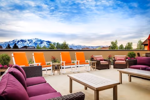 Outdoor patio area with purple cushioned wicker sofas, orange cushioned lounge chairs, wooden tables, and potted plants, set against a backdrop of trees and snow-capped mountains under a partly cloudy sky.