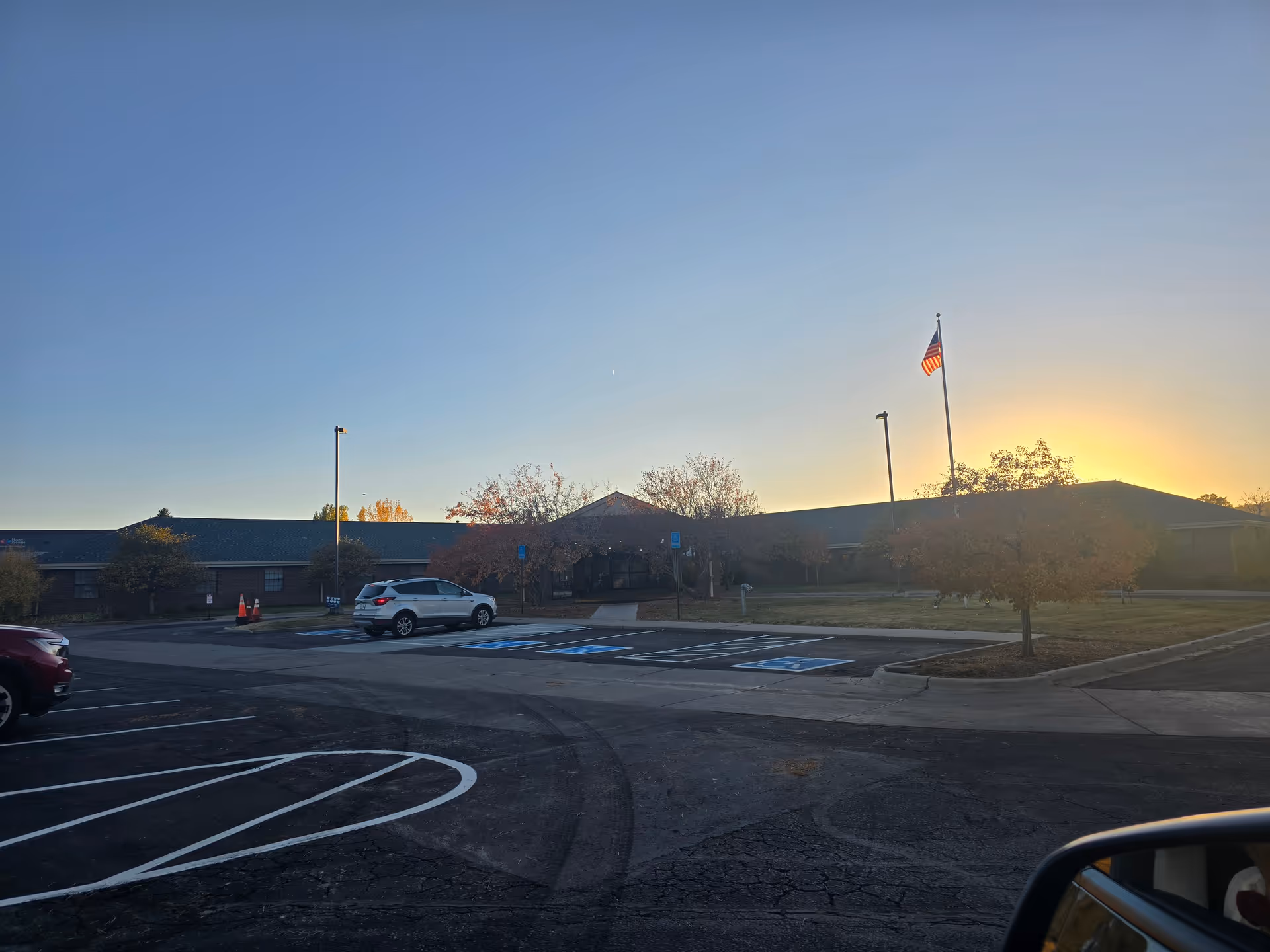 Exterior view of a single-story brick building with a parking lot in front, several parked cars, a few small trees, and an American flag on a flagpole at sunset.