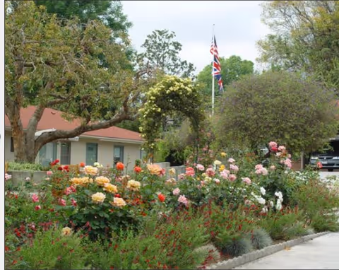 A garden area with various colorful blooming flowers and green shrubs in front of a single-story building with a red roof. There is a flagpole displaying the American and British flags, and trees surround the garden.