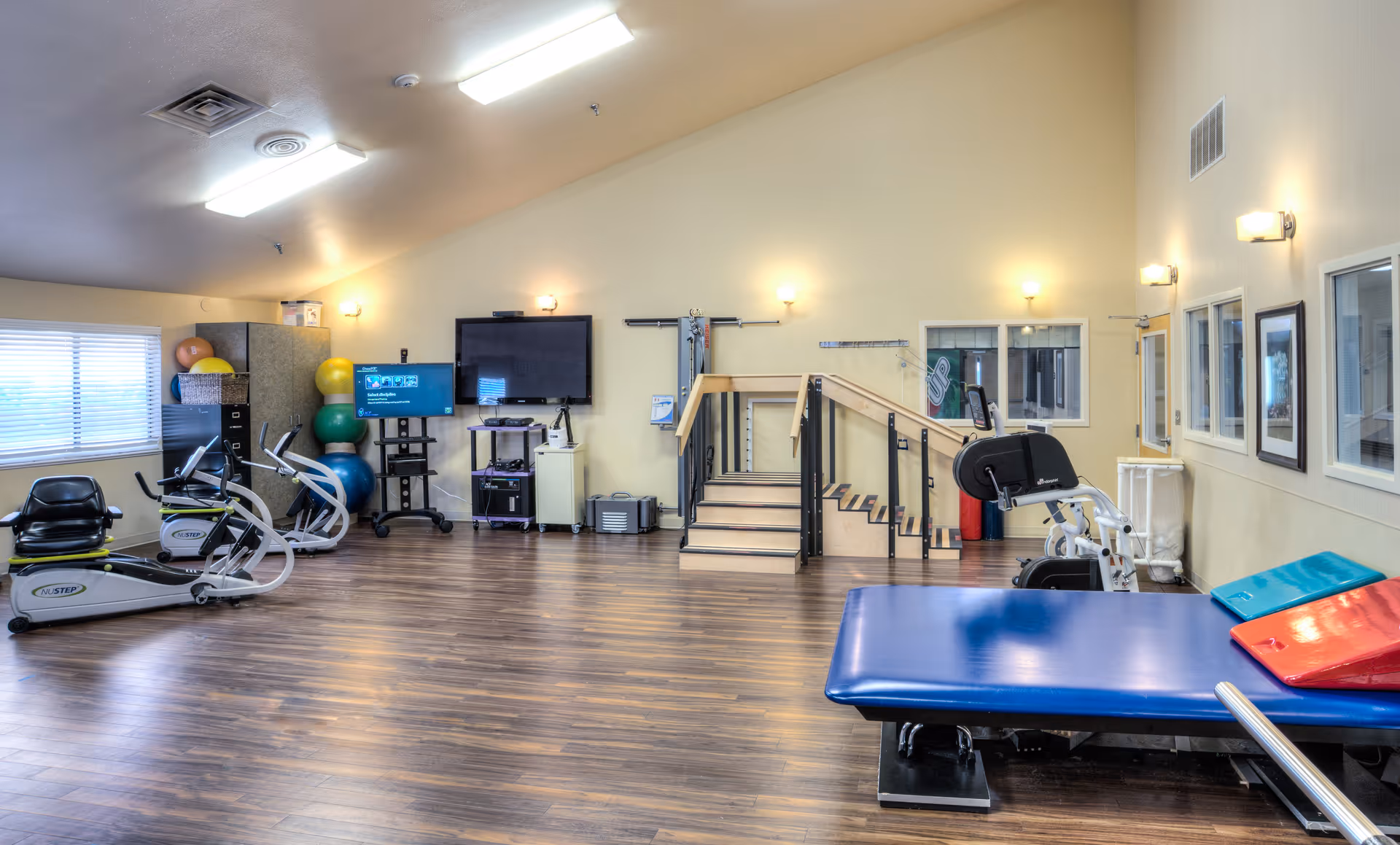 A spacious rehabilitation room with wooden flooring, exercise equipment including stationary bikes, therapy balls stacked in a corner, a blue therapy table with colorful wedges, a small staircase with handrails for physical therapy, and a large TV mounted on the wall. The room has several windows and bright overhead lighting.