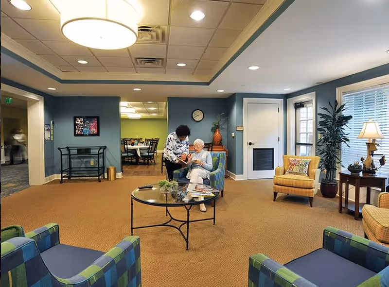 A cozy senior living facility common area with blue and yellow armchairs arranged around a glass coffee table. Two women, one standing and one seated, are interacting in the center of the room. The walls are painted blue with white trim, and there is a clock and decorative vase on a small table against the wall. A dining area is visible through an open doorway in the background.
