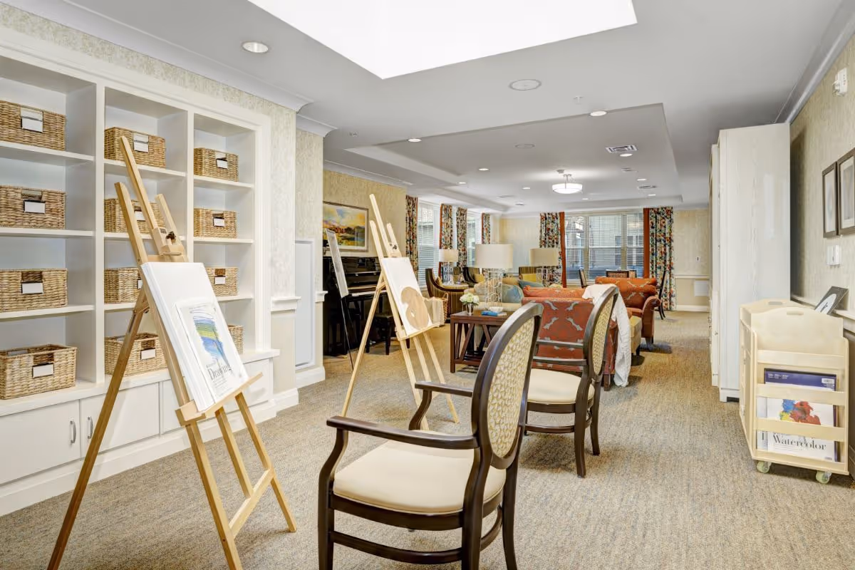 Well-lit communal activity room with easels and chairs in the foreground, shelving with wicker baskets to the left, and a seating area with sofas in the background.