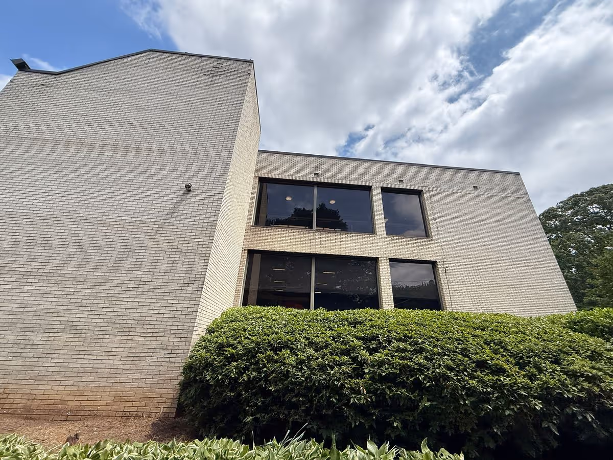 Brick exterior of a multi-story building with large windows, shrubs in front and a cloudy sky overhead.