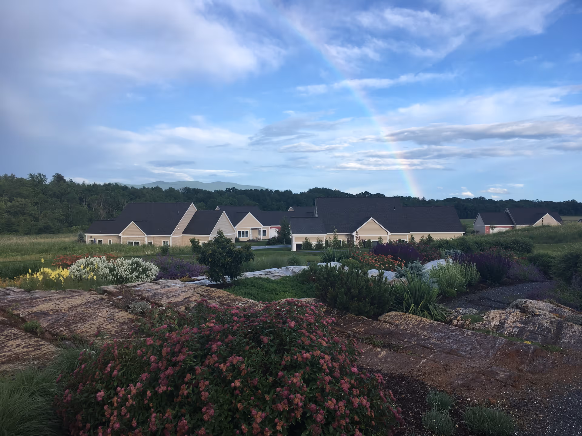 A scenic outdoor view of Eastview at Middlebury facility with beige buildings featuring dark roofs surrounded by lush greenery, colorful flowering plants, and large rocks in the foreground. A rainbow is visible in the partly cloudy blue sky above.