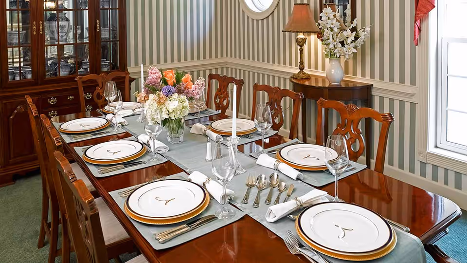 A formal dining room with a long polished wooden table set with plates, silverware, wine glasses and a floral centerpiece, surrounded by wooden chairs and a china cabinet.