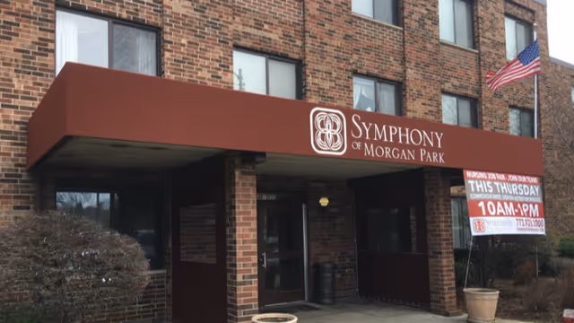 Exterior entrance of Symphony of Morgan Park facility with a maroon awning displaying the facility name. The building is made of brown brick, with several windows visible. An American flag is mounted on the right side near the entrance. A banner on the right side advertises a nursing job fair event.