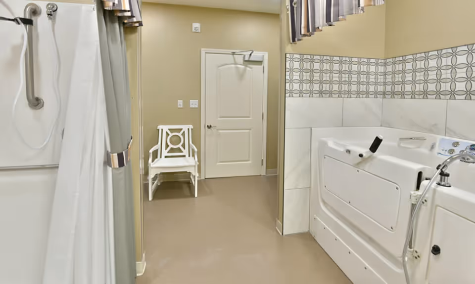 A senior-friendly bathroom featuring a walk-in bathtub with safety handles and a handheld showerhead. There is a white chair against the beige wall near a closed door, and the walls are decorated with patterned tiles above the bathtub. A shower curtain is partially visible on the left side.