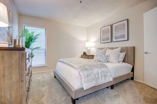 A cozy bedroom with a beige upholstered bed featuring white and patterned pillows, a soft gray blanket, and a wooden nightstand with a lamp and small plant. There is a wooden dresser with decorative items on top, a window with blinds, and two abstract framed artworks on the wall above the bed.