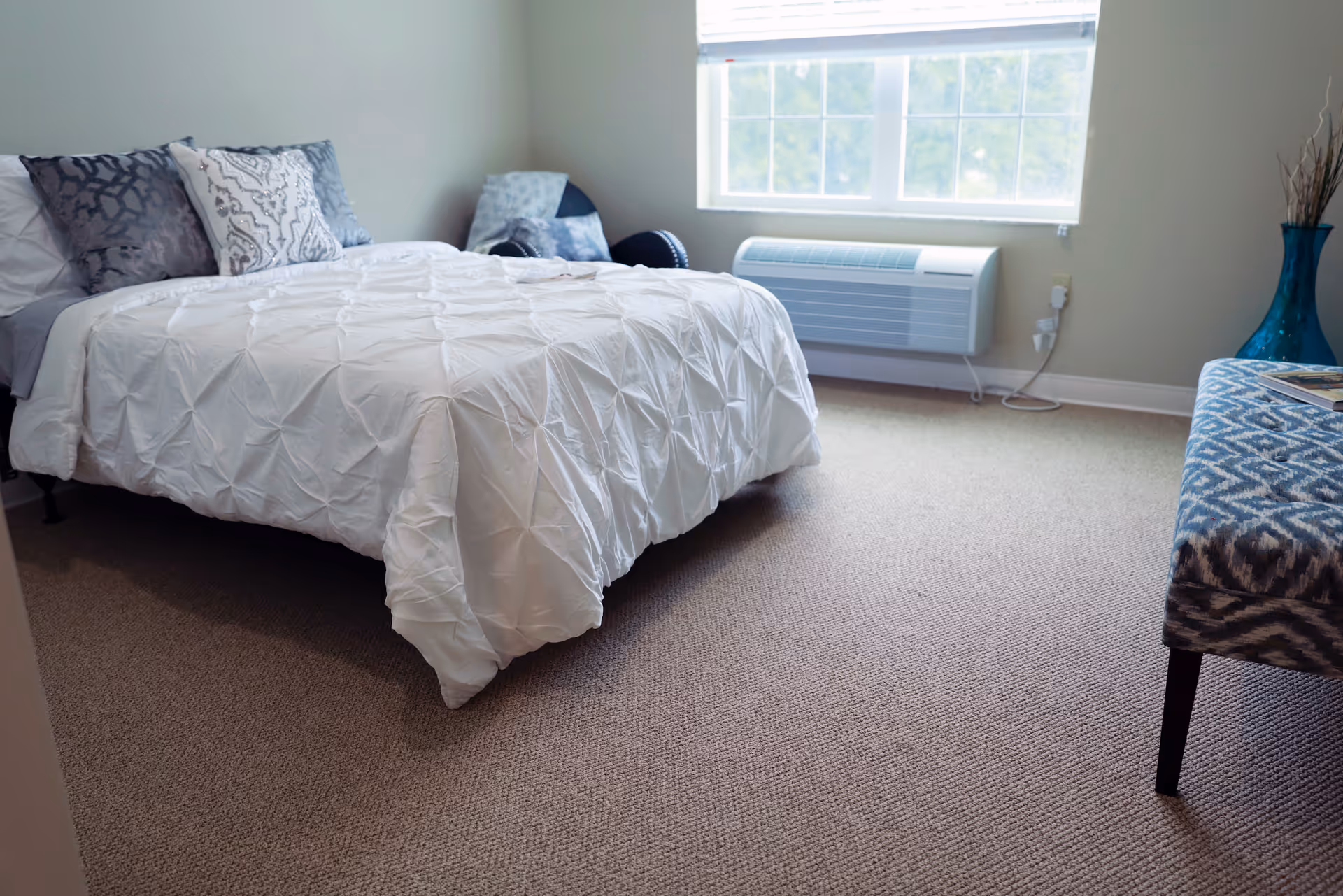 A bedroom with a neatly made bed covered in a white comforter and decorative pillows. There is a window with blinds letting in natural light, a blue patterned bench, and a blue vase with dried branches in the corner. The room has beige carpet flooring and light-colored walls.