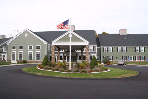 Two-story green building with a covered main entrance, circular driveway, landscaped island, and an American flag flying in front.