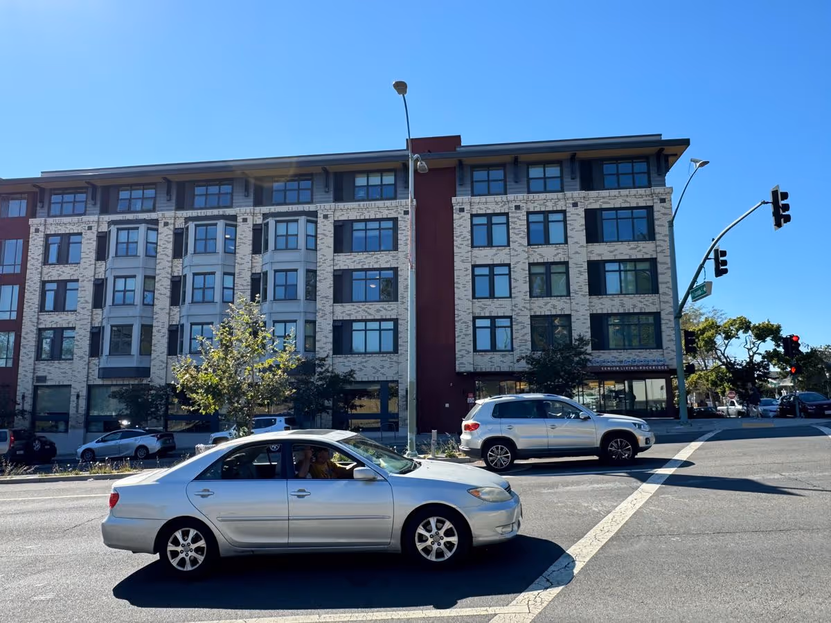 Five-story residential building with large windows facing a street intersection and cars stopped in the foreground.