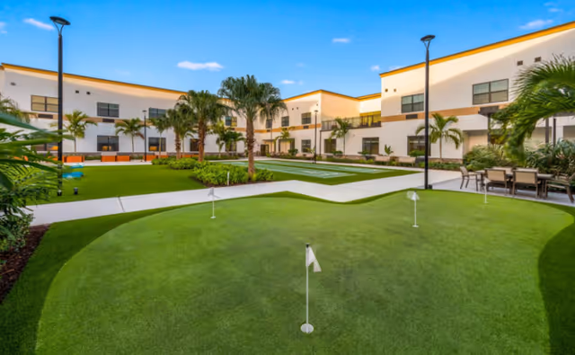 Sunny landscaped courtyard with a small putting green, palm trees, seating areas, and a surrounding two‑story building.