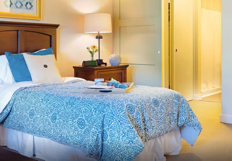 A cozy bedroom with a bed covered in a blue and white patterned bedspread, white and blue pillows, a wooden headboard, and a wooden nightstand with a lamp, a small vase with flowers, and an alarm clock showing 3:20. There is a tray with a cup and other items on the bed. In the background, a doorway leads to a bathroom with a shower curtain.