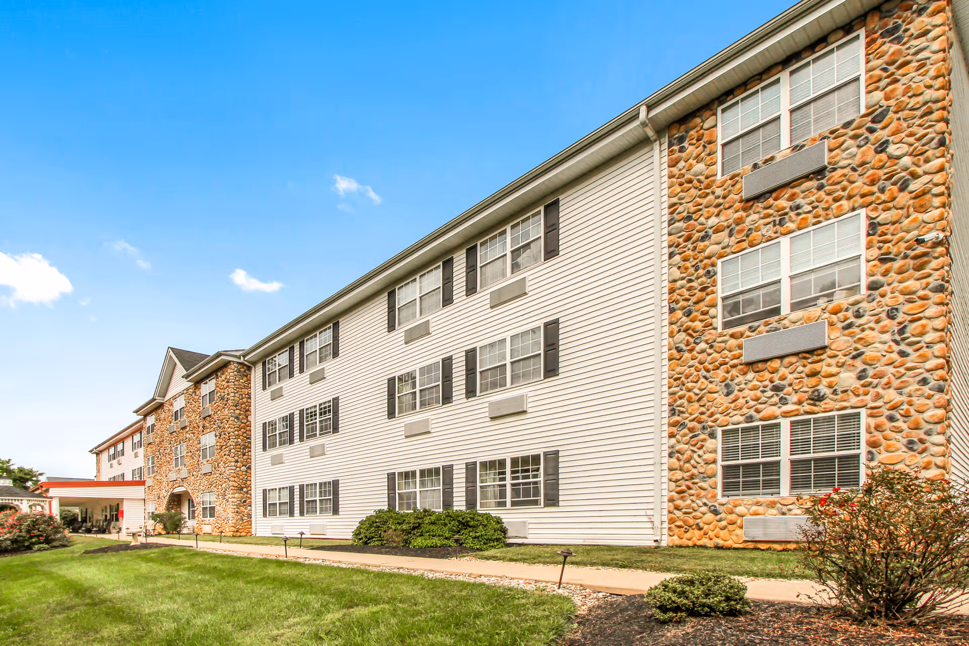 Front exterior of a three-story senior living building with stone and siding facade, lawn and walkway.