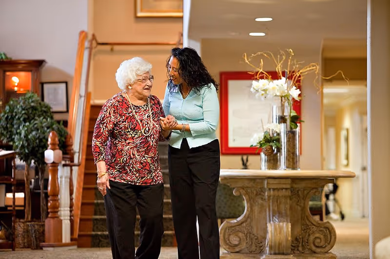 An elderly woman with white hair and glasses wearing a red patterned top and black pants is walking indoors, holding hands and smiling with a younger woman in a light blue sweater and black pants. They are in a warmly lit hallway with a decorative table holding flower arrangements and a staircase in the background.