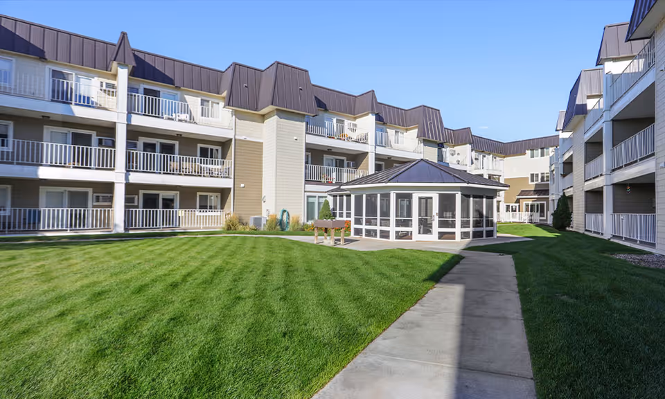 Outdoor courtyard area of Westpark Village senior living facility with well-maintained green grass, a paved walkway, and a central gazebo surrounded by multi-story residential buildings with balconies under a clear blue sky.