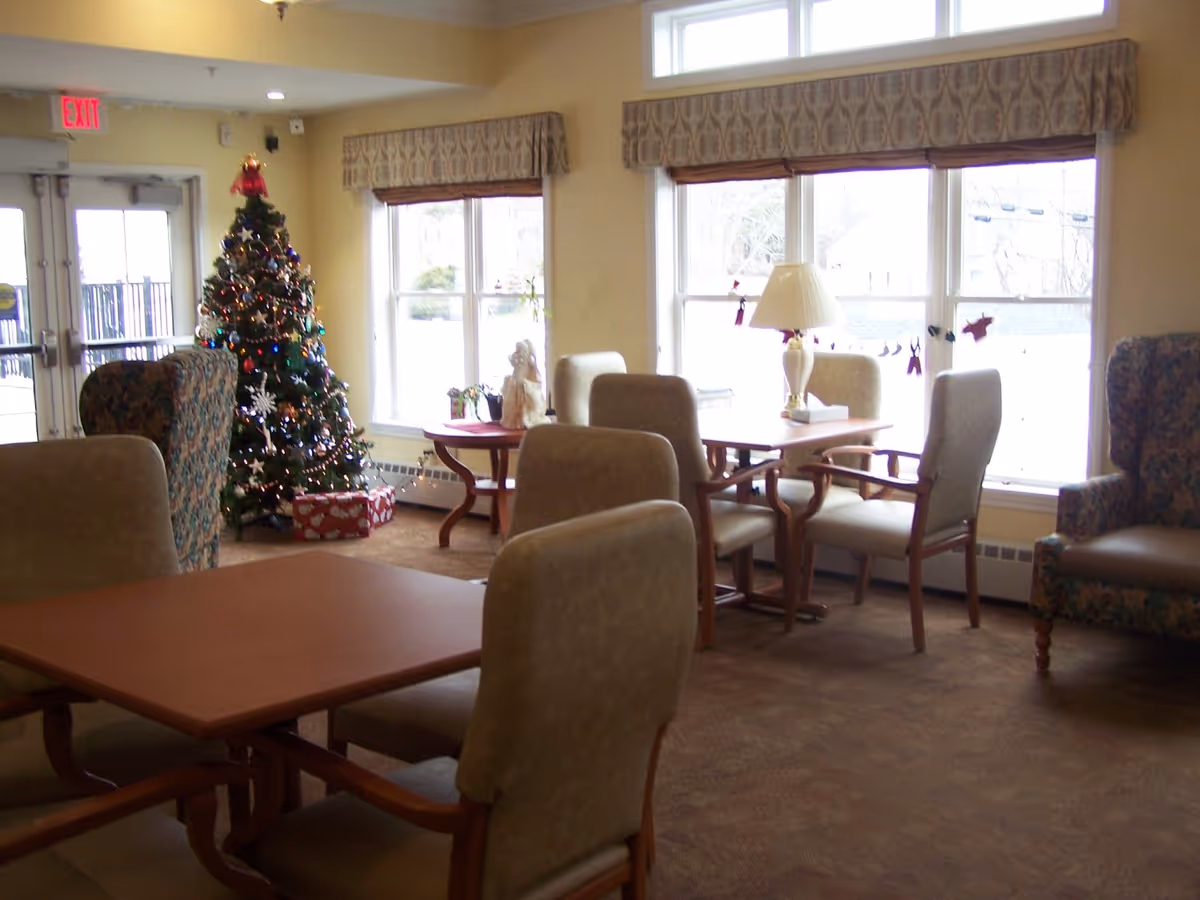 A cozy common area in Bayview Manor decorated for Christmas with a Christmas tree adorned with lights and ornaments. The room has several tables and chairs arranged for seating, large windows with patterned valances letting in natural light, and a table lamp on one of the tables. There is an exit door visible in the background.