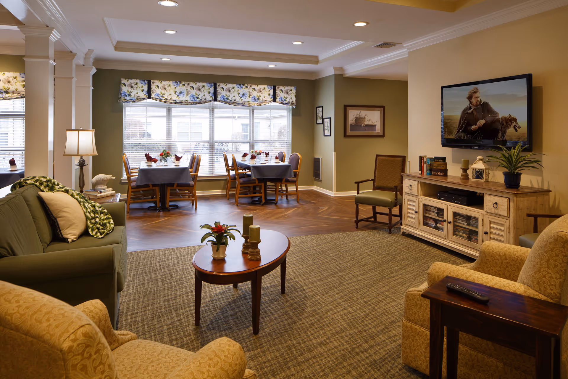 A cozy senior living common area with comfortable seating including a green sofa and beige armchairs, a wooden coffee table with decorative plants and candles, a TV mounted on the wall above a wooden cabinet, and a dining area with tables and chairs near large windows with floral valances.