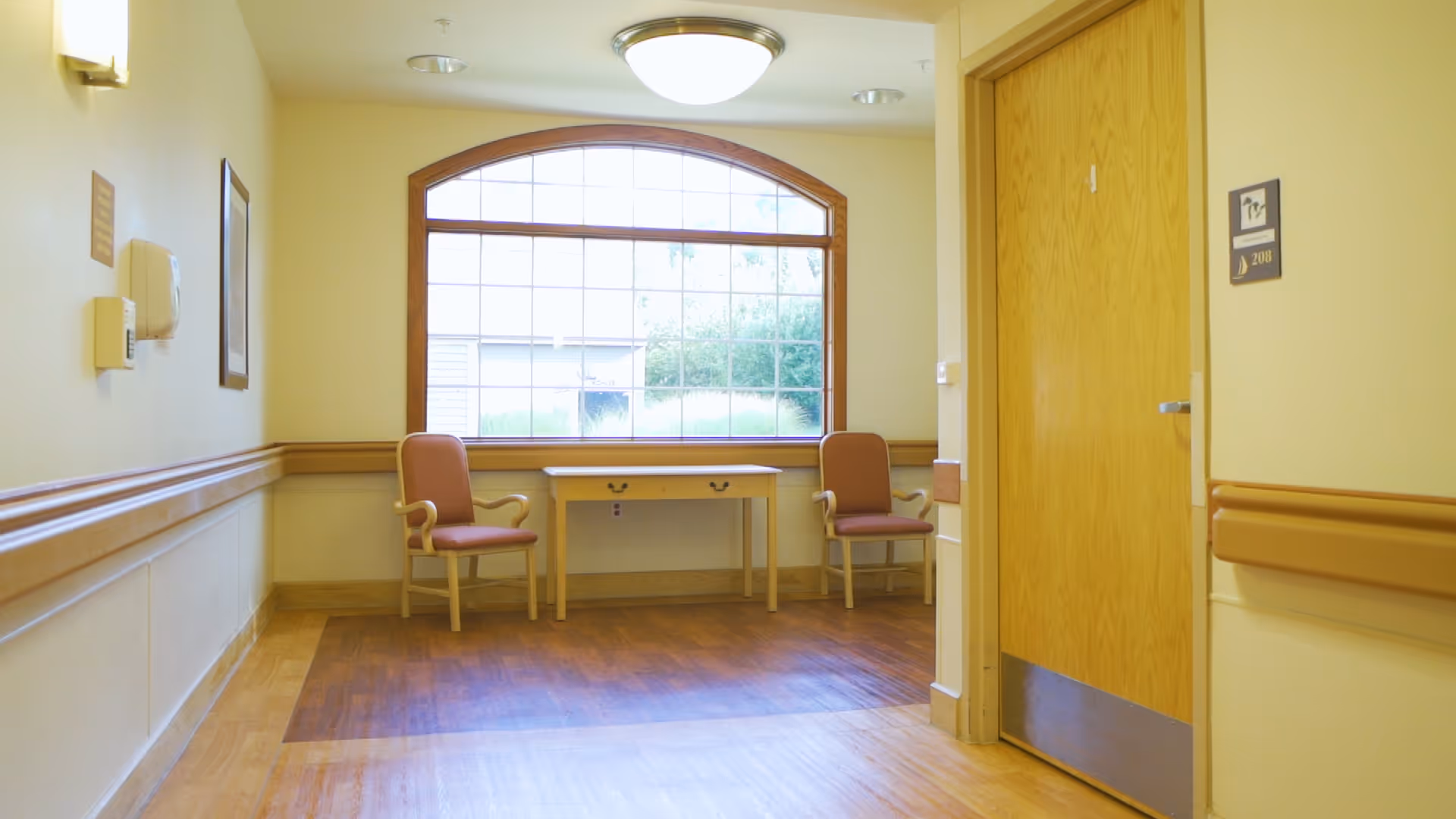 A quiet hallway area in a senior living facility with two wooden chairs with armrests and pink cushions placed on either side of a small wooden table. A large window with a wooden frame lets in natural light, illuminating the light yellow walls and wooden floor. A closed wooden door with the room number 208 is visible on the right side of the image.