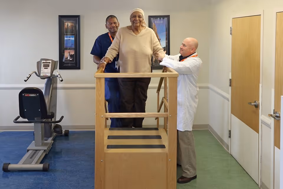 An elderly woman is assisted by two healthcare professionals as she practices walking on a wooden rehabilitation step platform in a therapy room with exercise equipment and motivational posters on the wall.