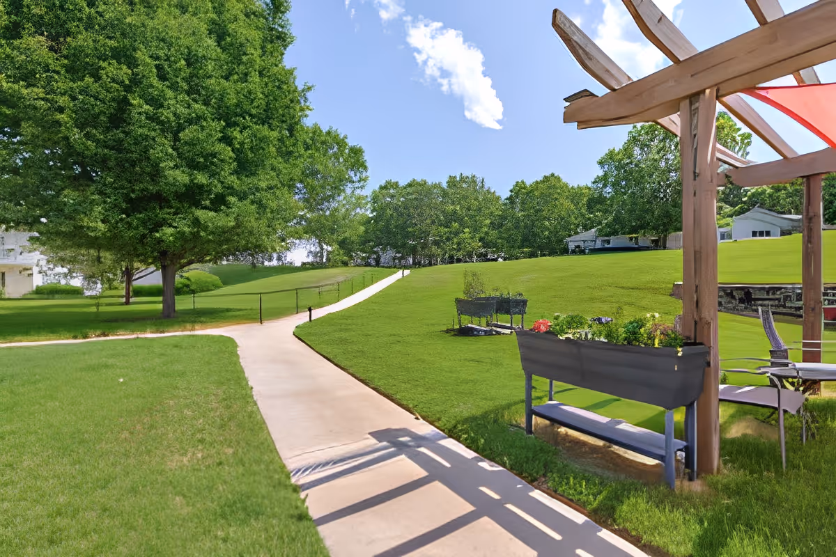 Sunlit outdoor walking path through a grassy lawn with a pergola, bench planters, and trees in the facility grounds.
