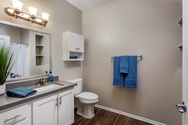 A clean bathroom with a white vanity cabinet, gray countertop, and a sink. Above the sink is a large mirror with a three-light fixture. There is a white toilet next to the vanity, a small white wall cabinet above the toilet, and a towel rack on the wall holding two blue towels. The floor is wood-style and the walls are painted light beige.