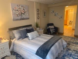 A cozy bedroom with a neatly made bed featuring striped bedding and multiple pillows. There is a white nightstand with a lamp and decorative items on the left side of the bed. A dark armchair with a throw blanket is positioned in the corner near the doorway. The walls are decorated with floral artwork, and a patterned rug covers the floor.