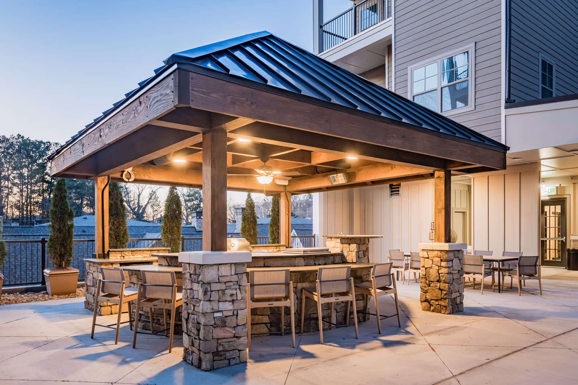 Outdoor covered seating area with stone pillars and a metal roof at a senior living facility. The area includes a bar-style counter with chairs and additional tables and chairs nearby. Trees and building exteriors are visible in the background during dusk.