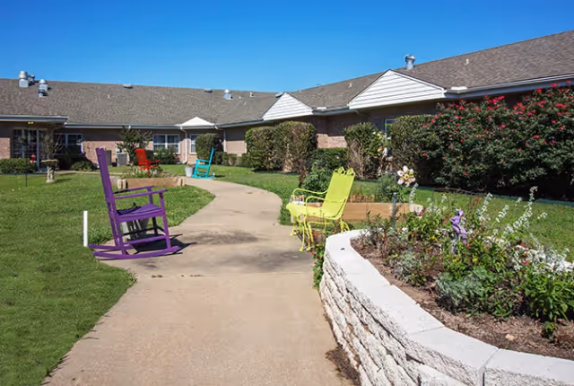 Outdoor courtyard area at Bluebonnet Court featuring a paved walkway, colorful rocking chairs in purple, yellow, red, and blue, green grass, flower beds with blooming plants, and a brick building with windows under a clear blue sky.
