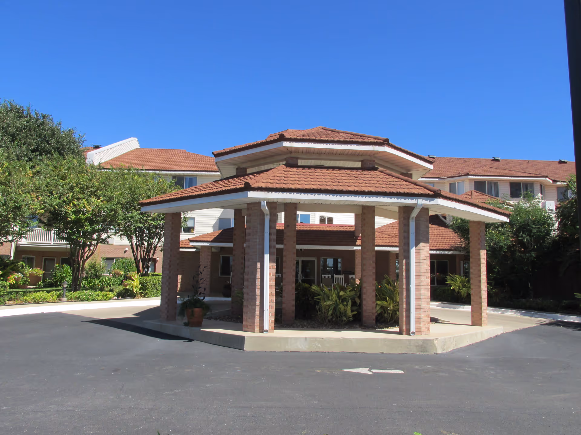 Exterior view of a retirement community building with a covered entrance supported by brick pillars, surrounded by greenery and trees under a clear blue sky.