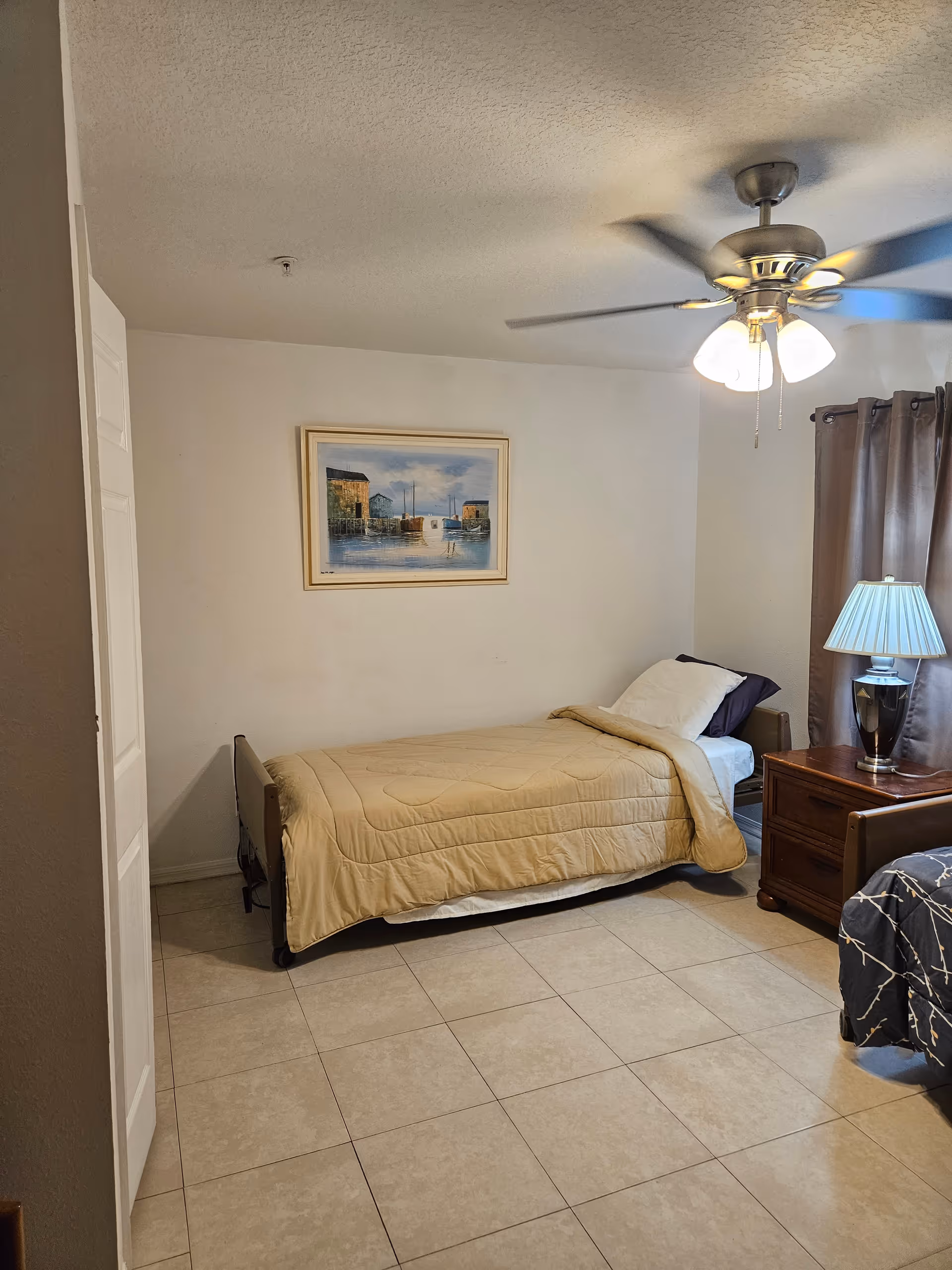 A small assisted-living bedroom with a single bed, wooden nightstand and lamp, ceiling fan, framed artwork, and tiled floor.