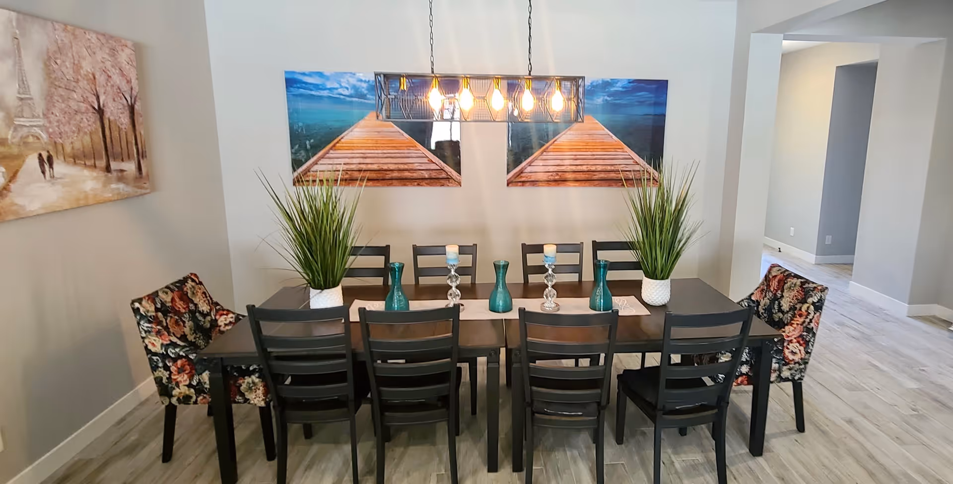 A modern dining room with a long dark wooden table surrounded by eight chairs, two of which have floral upholstery. The table is decorated with two potted green plants, three blue vases, and two candle holders. Above the table hangs a rectangular light fixture with exposed bulbs. On the wall behind the table are two large paintings depicting a wooden pier extending into a body of water. The room has light-colored walls and flooring, with an open doorway visible on the right.