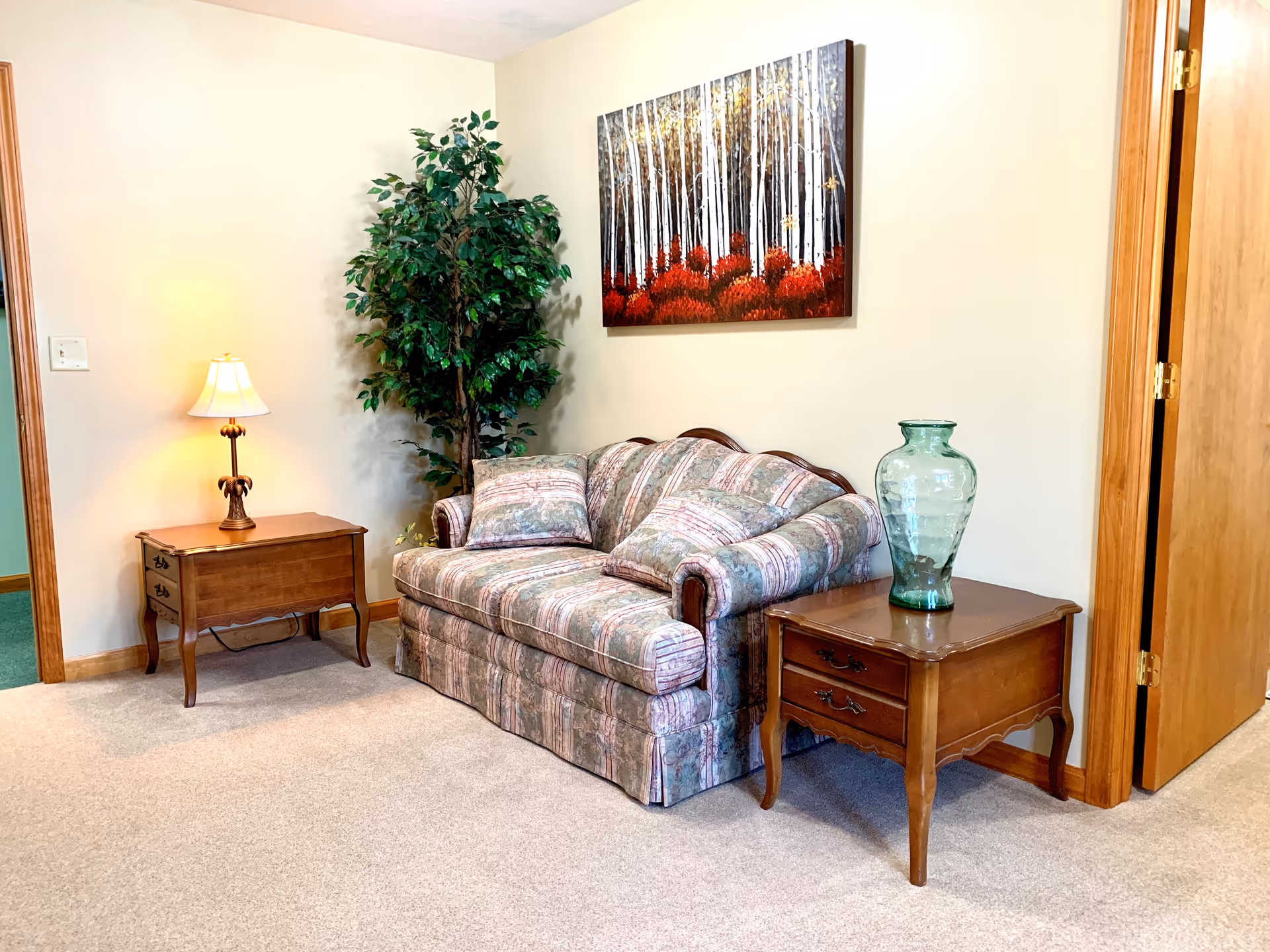A small sitting area with a patterned loveseat between two wooden side tables, a lamp, a tall potted plant, and a framed forest painting on the wall.
