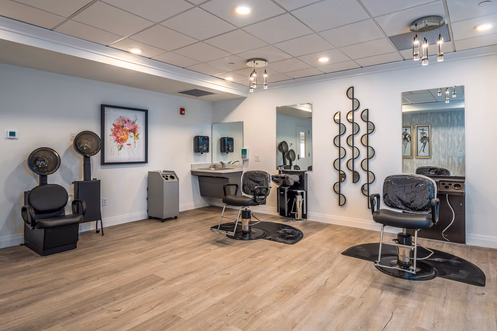Interior view of a salon area in a senior living facility with two black salon chairs in front of large mirrors, two hair drying stations, a sink with a countertop, modern light fixtures on the ceiling, and decorative wall art.