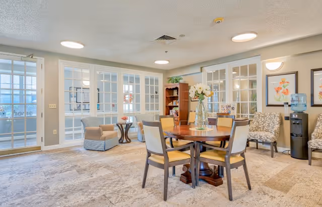 A bright and spacious common area with a round wooden table and four chairs in the center. A vase with white flowers is placed on the table. The room has beige walls and carpeted floors. There are two patterned armchairs and a water dispenser against the wall, along with framed floral artwork. Glass-paneled doors and windows separate this room from an adjacent area, and a small side table with a chair is visible near the doors.