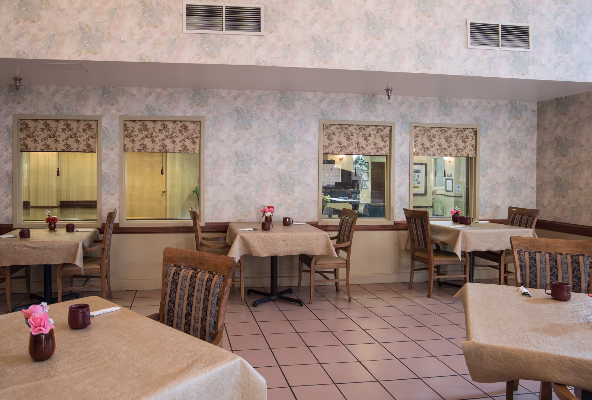 Interior view of a dining room with several tables covered in beige tablecloths, each set with a small vase of pink flowers and a maroon cup. The room has tiled floors, floral patterned window shades, and light-colored walls with a textured finish.