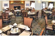 A spacious dining room with multiple tables and chairs arranged neatly. Each table is set with white cups, saucers, and silverware. The room features a patterned carpet, a brick fireplace with a mantel decorated with plants and framed pictures on the walls. Large windows with curtains allow natural light to brighten the space.